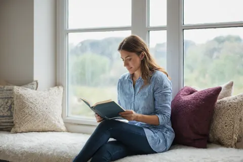 A woman reading a book on a window seat with pillows and natural light