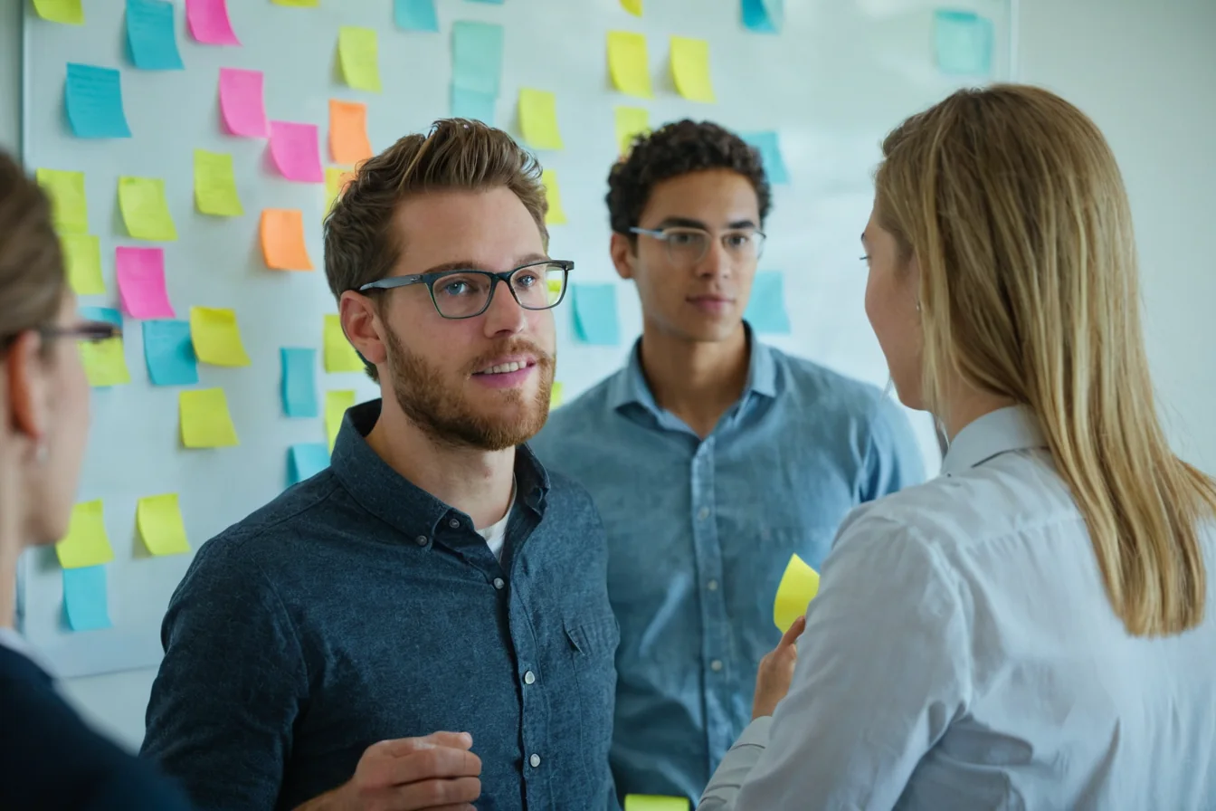 Four people in an office meeting discussing ideas on a whiteboard with colorful sticky notes.
