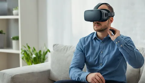 Man wearing VR headset and holding controller while seated on gray couch in modern living room