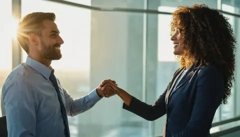 Two business colleagues shaking hands in a bright office with sunlight streaming through windows.