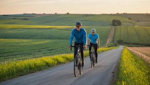 Two men cycling on a country road surrounded by green fields and yellow flowers at sunset