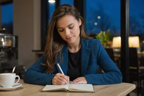 Woman writing in notebook at cafe table with coffee cup nearby