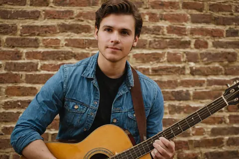 Young man holding acoustic guitar in front of brick wall